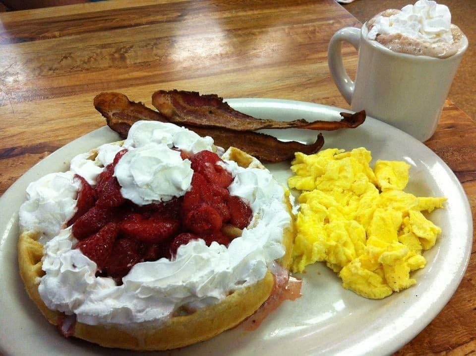 Waffle piled with strawberries and whipped cream, bacon, scrambled eggs and a mug of whipped-cream cocoa
